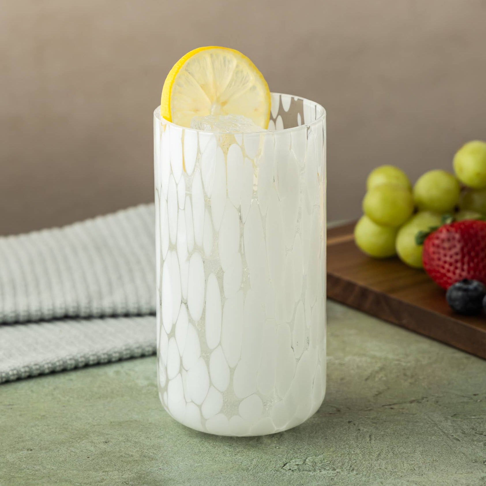 A tall, textured white glass tumbler with a dripping marble pattern, filled with a white beverage and garnished with a lemon slice, set on a stone tabletop with a striped cloth and fresh fruit in the background.