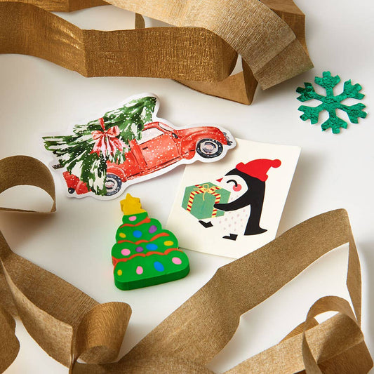 A set of Christmas-themed stickers or decals arranged on a white surface with decorative gold tissue paper. The stickers include a red car carrying a green Christmas tree, a green Christmas tree with colorful ornaments, a penguin in a red Santa hat holding a gift, and a green snowflake.