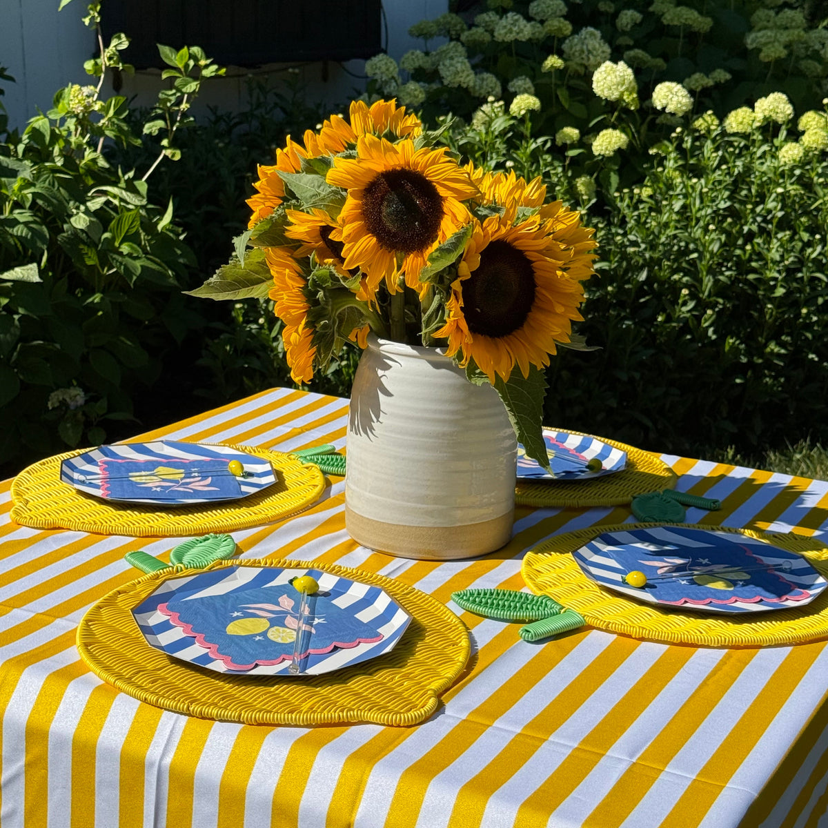 A styled outdoor dining table setup with a yellow and white striped tablecloth, matching placemats, woven yellow placemats, blue patterned plates, and a white vase filled with sunflowers, set in a garden environment.
